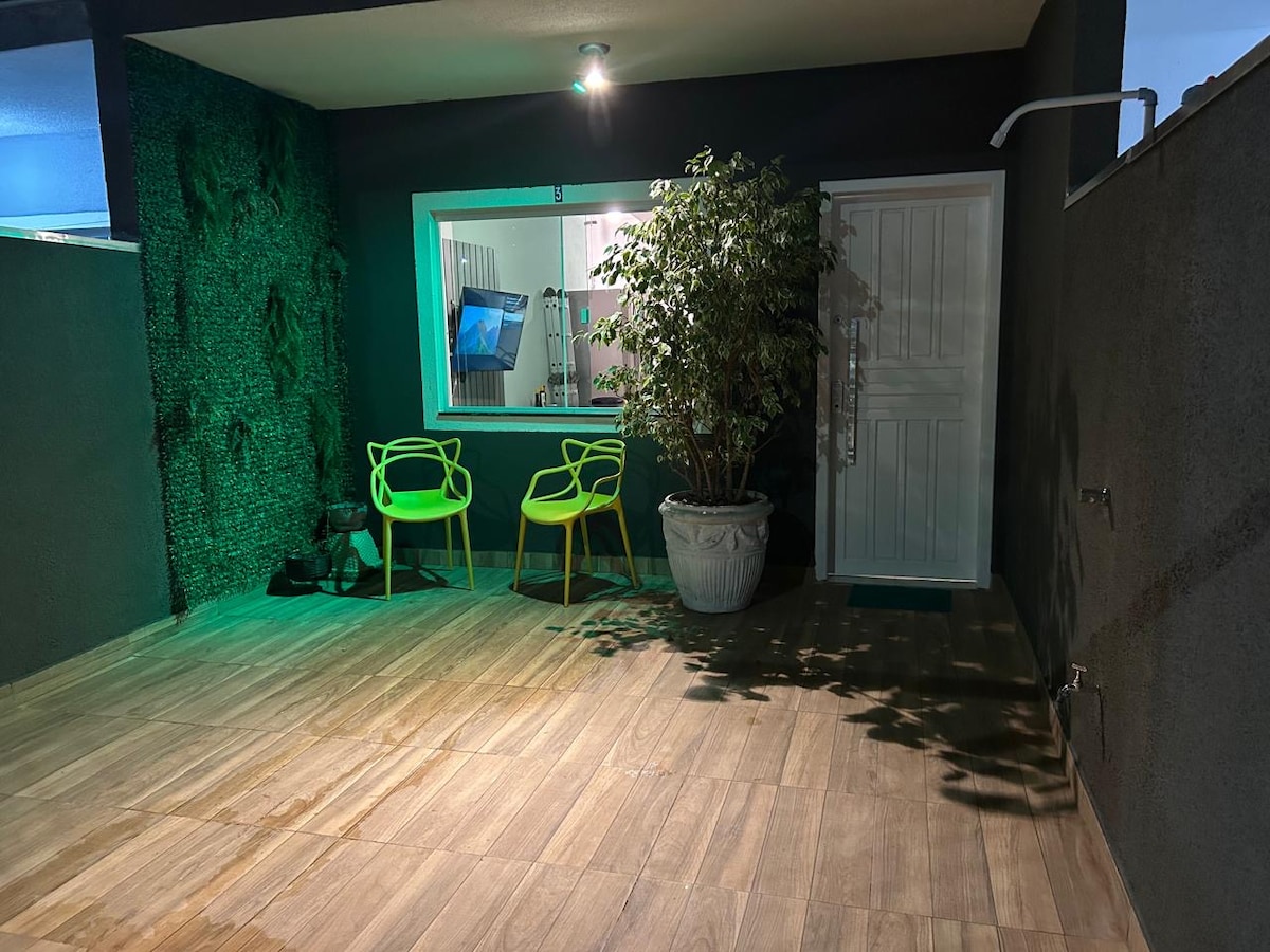 An outdoor area features a wooden deck, complemented by a green plant in a large pot. Two bright green chairs are positioned nearby, creating a welcoming space. Soft lighting enhances the ambiance, while the entrance door is seen in the background.