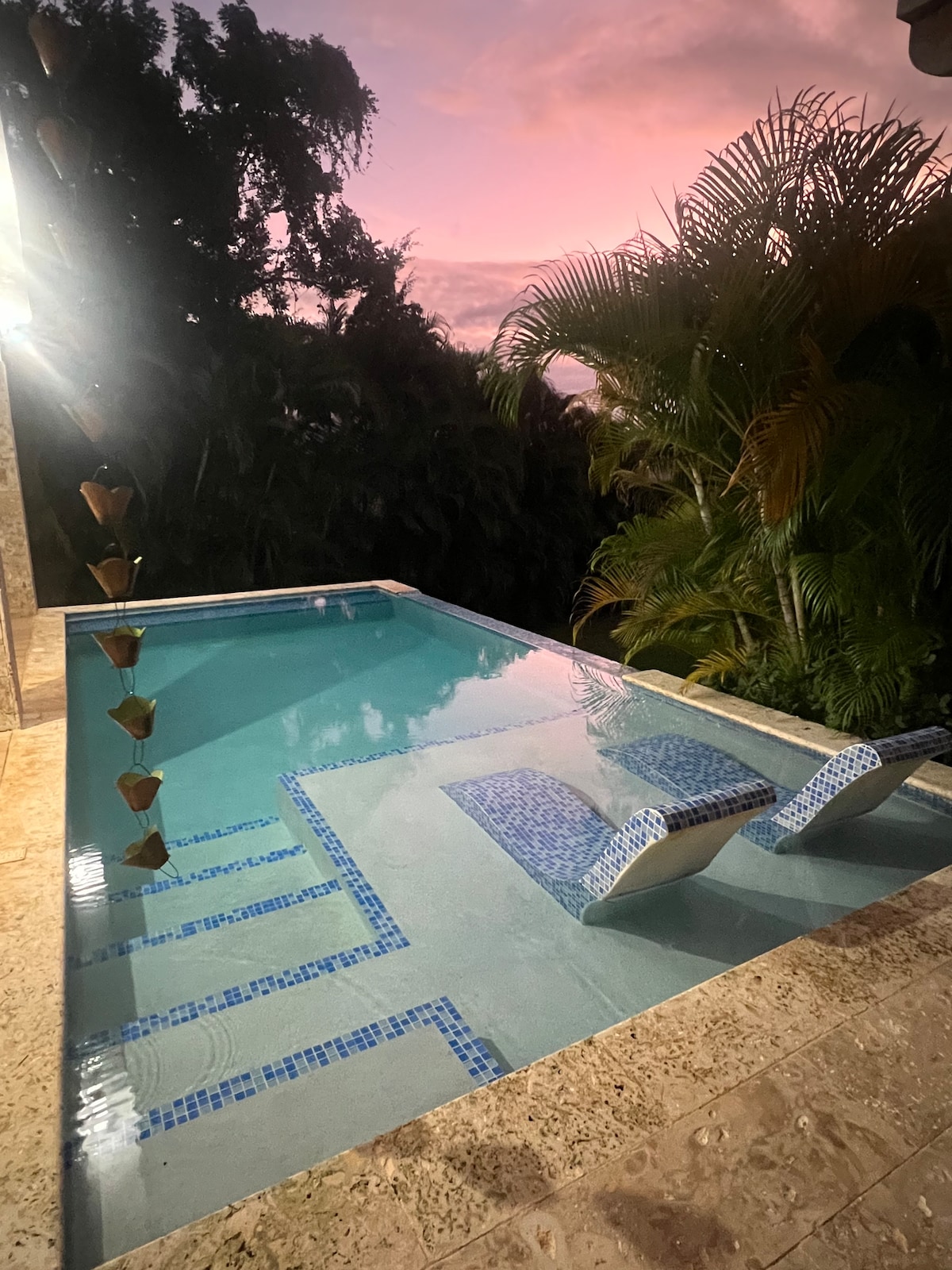 A serene swimming pool is surrounded by lush greenery, with two comfortable lounge chairs partially submerged in the water. The setting is enhanced by soft evening light and a sky painted in warm shades, creating a peaceful outdoor retreat.