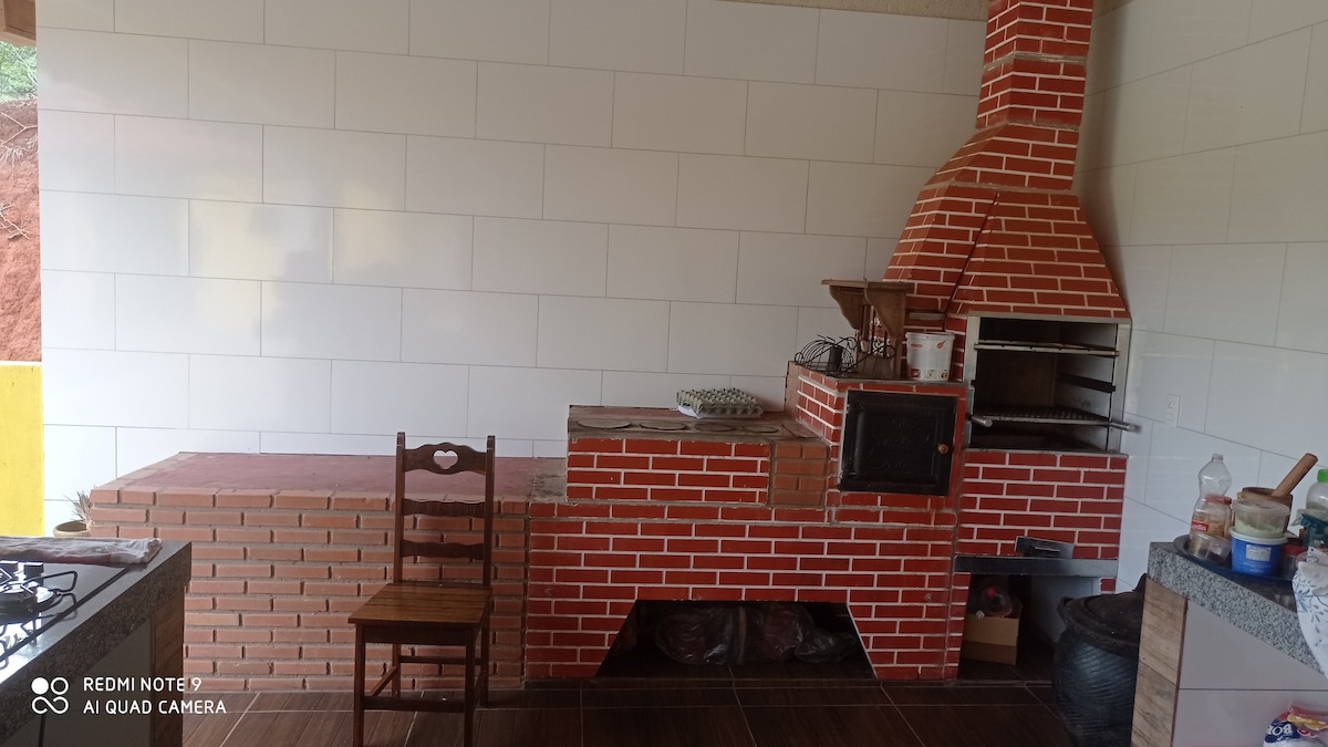 An outdoor kitchen area features a brick grill setup with a rustic chimney. A single wooden chair is positioned beside a long counter made of brick. The walls are tiled in white, and various cooking utensils and containers are neatly arranged on the countertop.