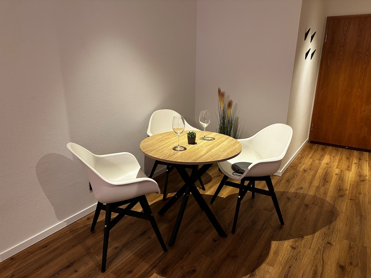A small dining area is depicted with a round wooden table surrounded by three white chairs. Two wine glasses rest on the table, and a small plant adds a touch of greenery. The warm wood floor complements the soft white walls.
