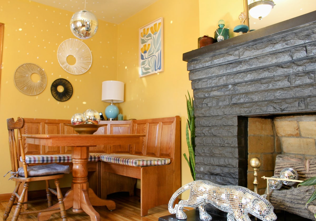 A dining area is showcased with a wooden table surrounded by several chairs. The yellow walls are complemented by decorative wall art. A fireplace is visible with a metallic sculpture positioned nearby, while disco balls reflect light around the space, creating an inviting ambiance.