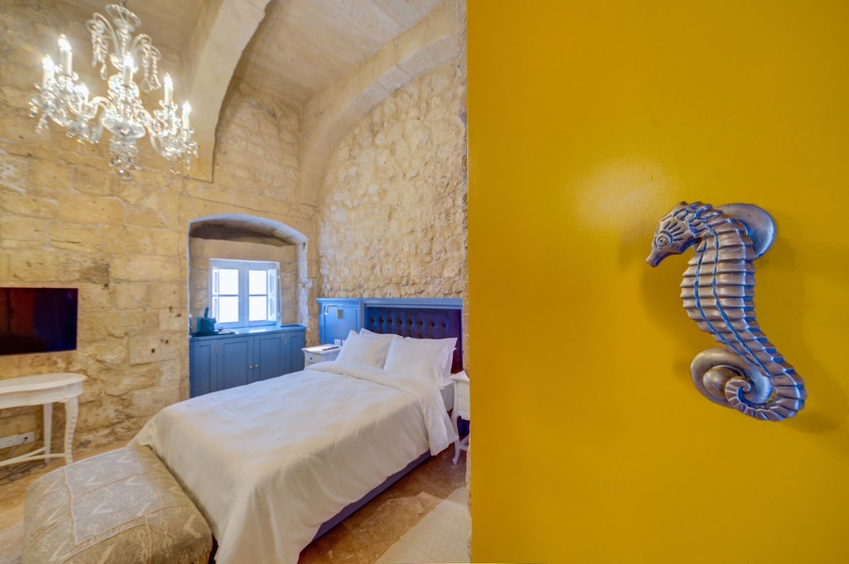 A bedroom is illuminated by a chandelier, featuring a stone wall and a vibrant yellow accent wall. A spacious bed is dressed in white linens, while a small TV is mounted on the wall. A decorative seahorse door handle enhances the room's charm.