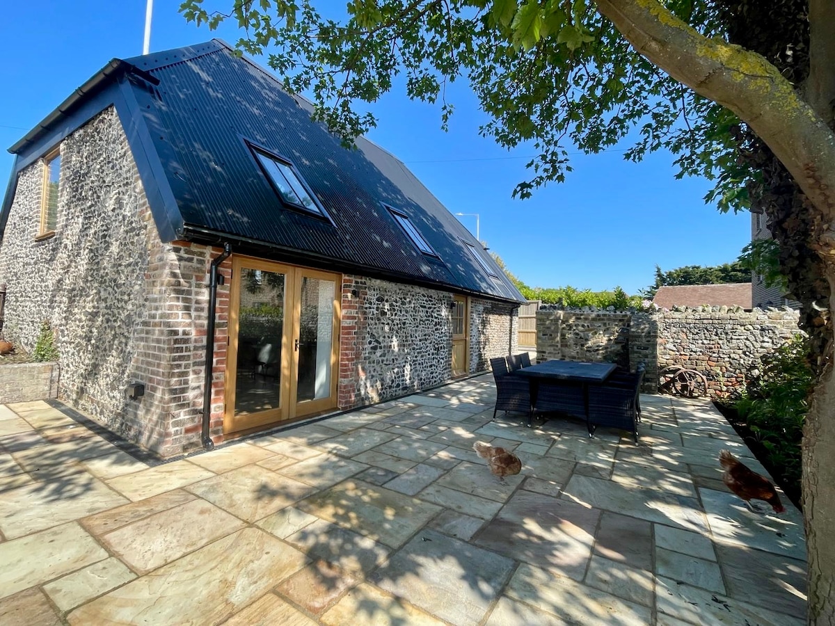 A charming outdoor patio area features a stone surface and a set of black wicker dining furniture. The barn's textured stone and slate exterior is visible, along with free-roaming chickens exploring the space. Lush greenery and clear blue skies create a tranquil setting.