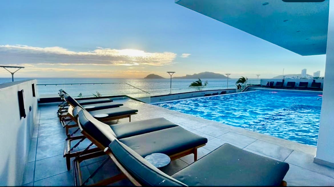 A tranquil pool area is highlighted by lounge chairs with ocean views. The spacious pool reflects the sky, with gentle waves visible in the distance. Sunlight casts a warm glow over the scene, creating an inviting atmosphere for relaxation.