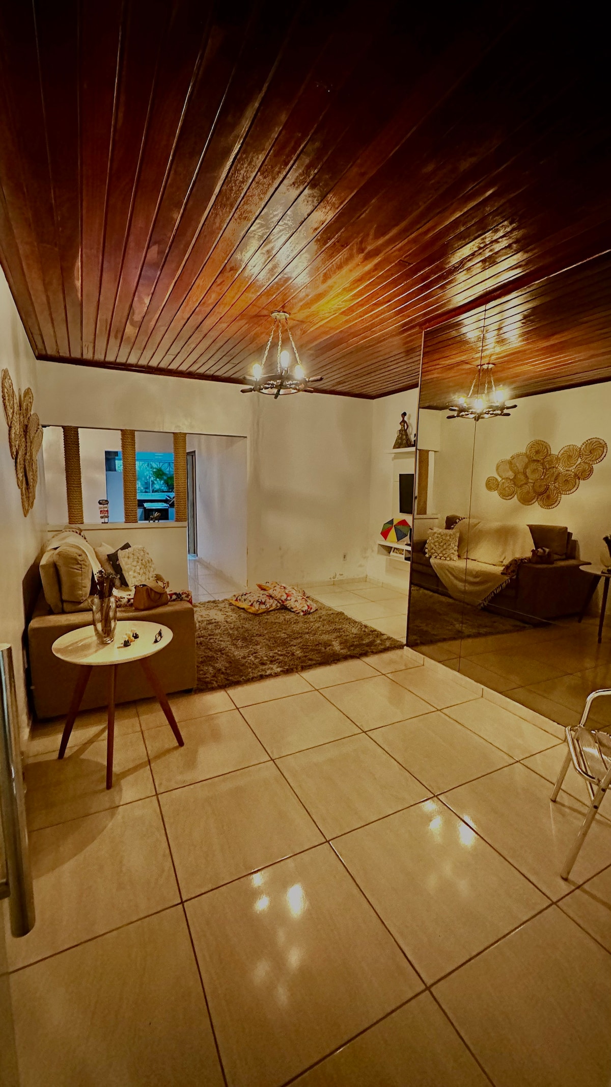 A spacious living area features polished tile flooring and a wooden ceiling. A cozy seating arrangement is present, with decorative wall art and soft textures adding warmth. Natural light enters through large windows, creating a bright and inviting space for relaxation.