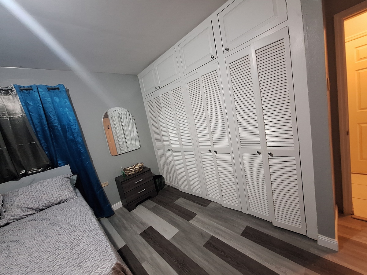 A sleeping area is featured with a spacious bed dressed in neutral tones. Large white cabinetry with shuttered doors provides ample storage. A full-length mirror is mounted on the wall, and a blue curtain filters natural light from a nearby window.