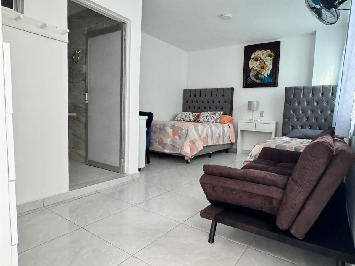 A bright and airy room features two beds with patterned bedding, accompanied by a sleek nightstand and lamp. A comfortable brown chair adds seating, while a bathroom door is visible at the back. Natural light enters through a window, enhancing the spacious feel.