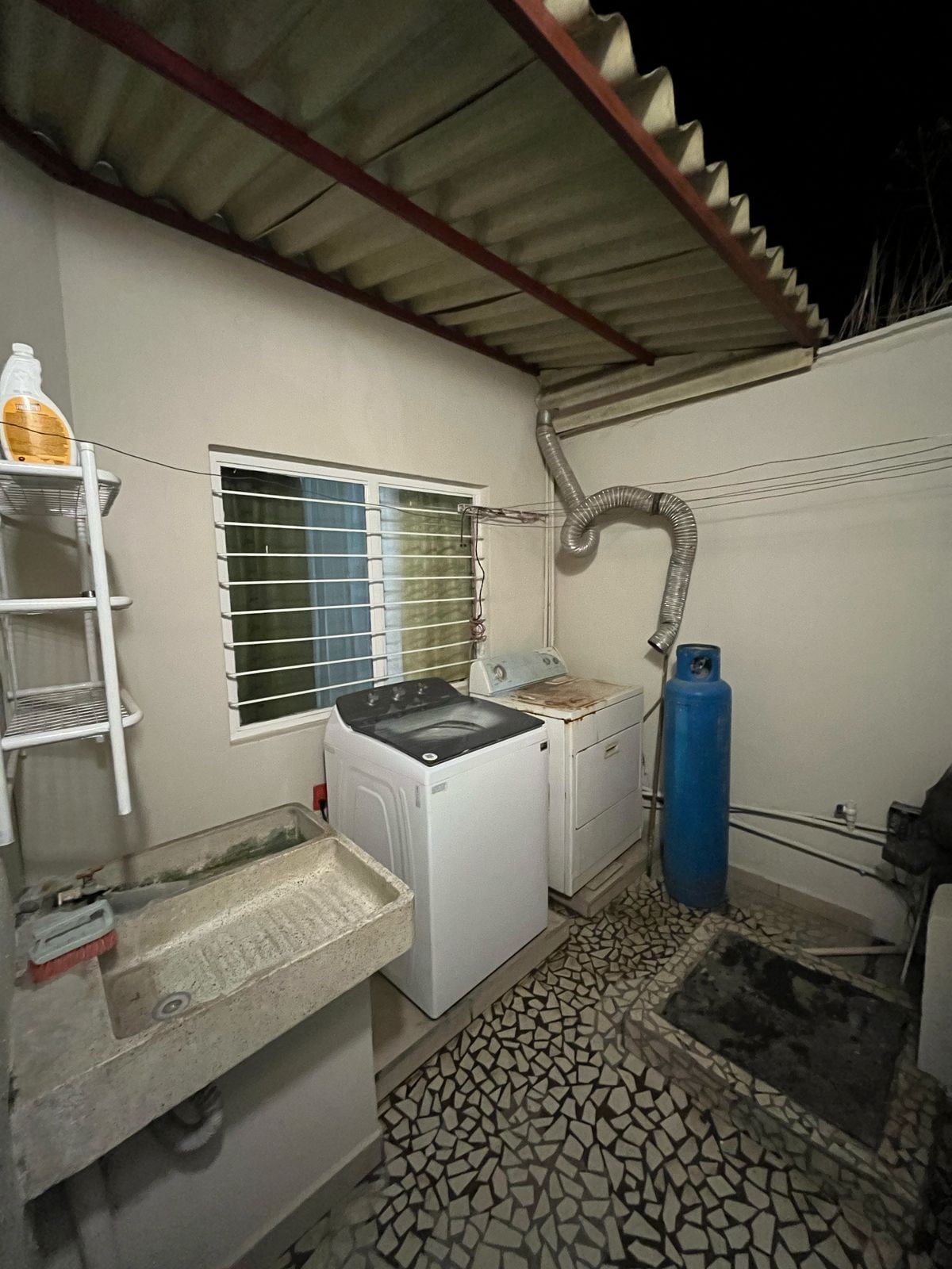 A functional laundry area is visible, featuring a washing machine and a drying space. A utility sink sits on one side, while a blue gas cylinder is positioned in the corner. Light enters through a small window, with mosaic flooring beneath.
