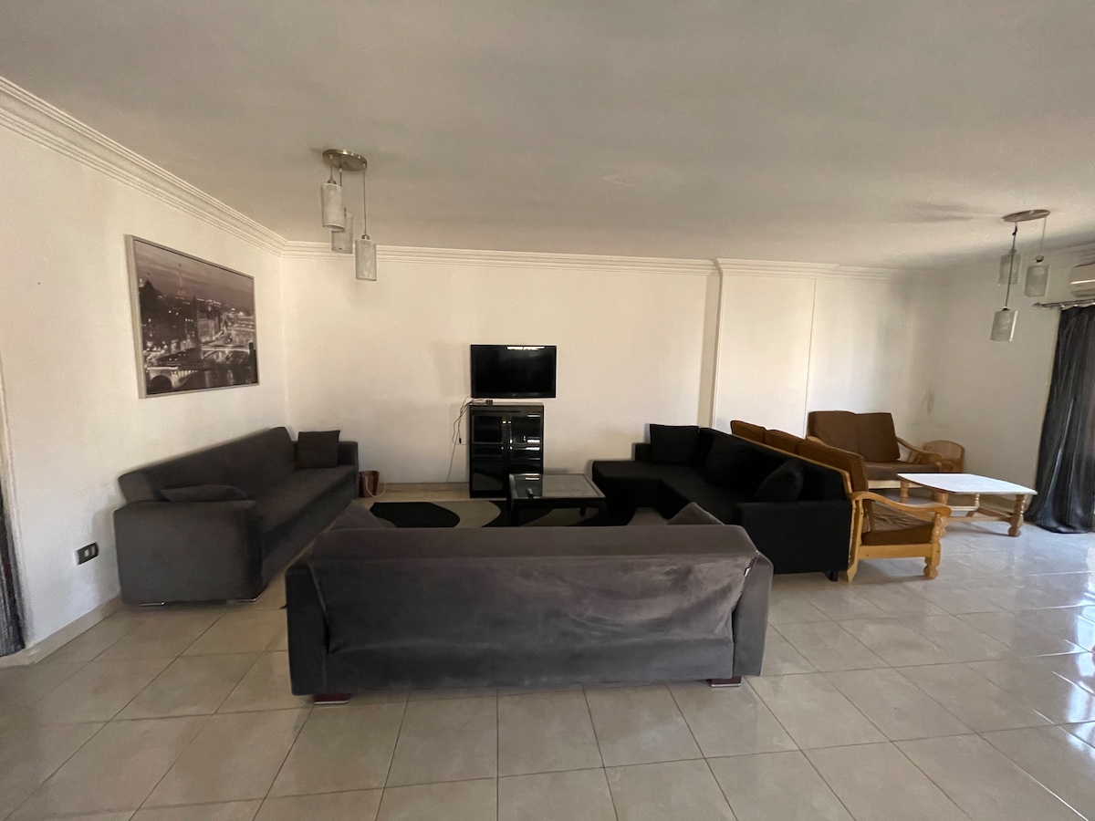 A spacious living area is equipped with multiple seating options, including fabric sofas and a wooden chair. A television is positioned on a stand against a wall. Large windows allow natural light to fill the space, with light-colored tiles covering the floor.