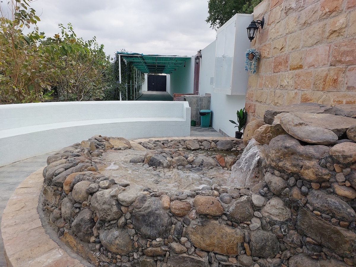 A stone fountain, featuring smooth pebbles and clear water gently flowing, is situated in a serene outdoor space. Surrounding greenery enhances the natural ambiance, while a light-colored wall adds contrast to the setting.