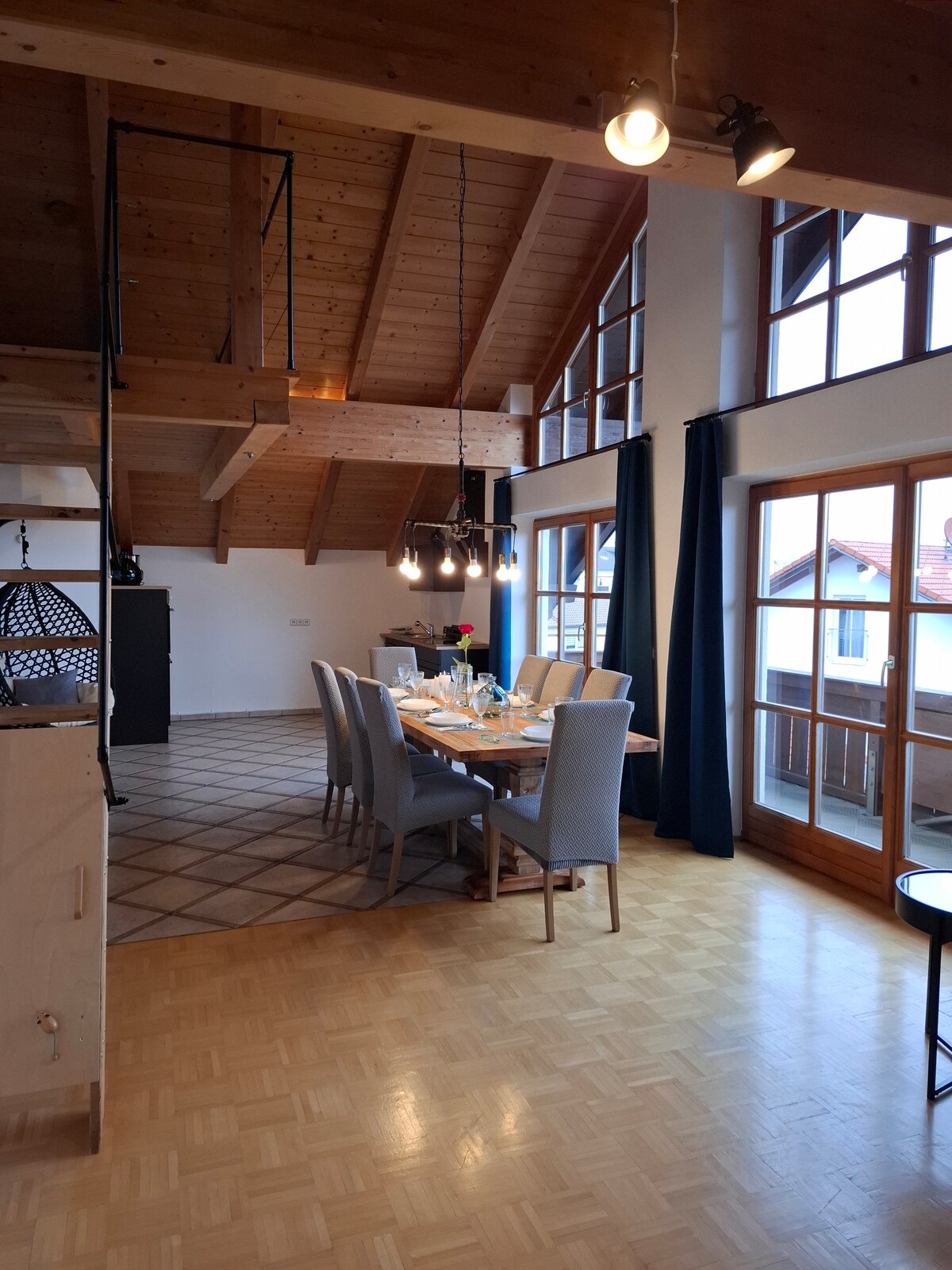A spacious dining area is showcased, featuring a large wooden table surrounded by six grey upholstered chairs. High ceilings with wooden beams create an airy feel. Large windows allow natural light to fill the room, while a dark accent wall adds depth. Elegant lighting fixtures complement the decor.