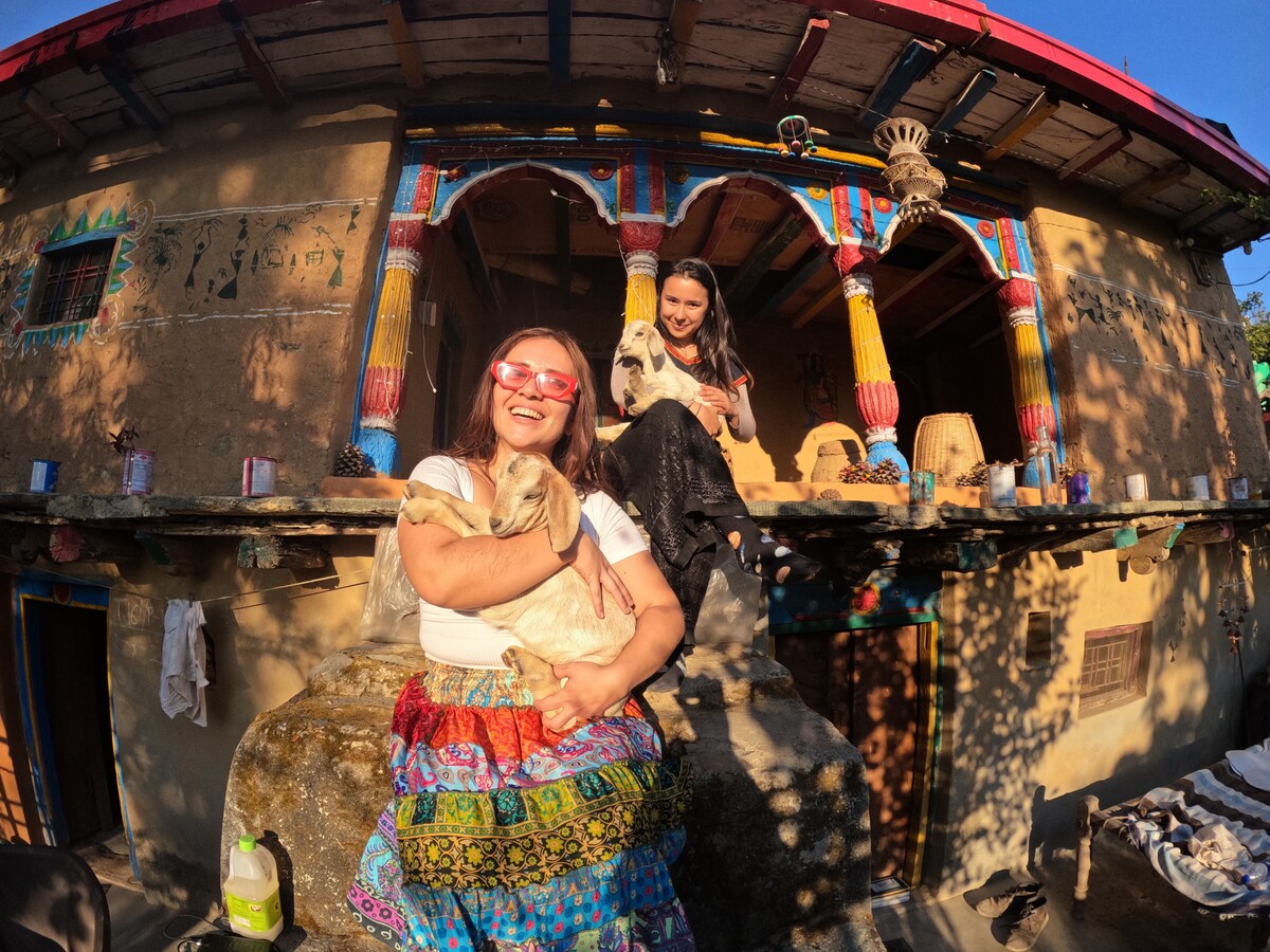 A woman dressed in colorful attire holds a goat while sitting on a large rock outside a traditional stone home. The home features vibrant wooden beams and arches, with sunlight creating soft shadows on the rustic façade. Two other goats are perched on the railing beside her.
