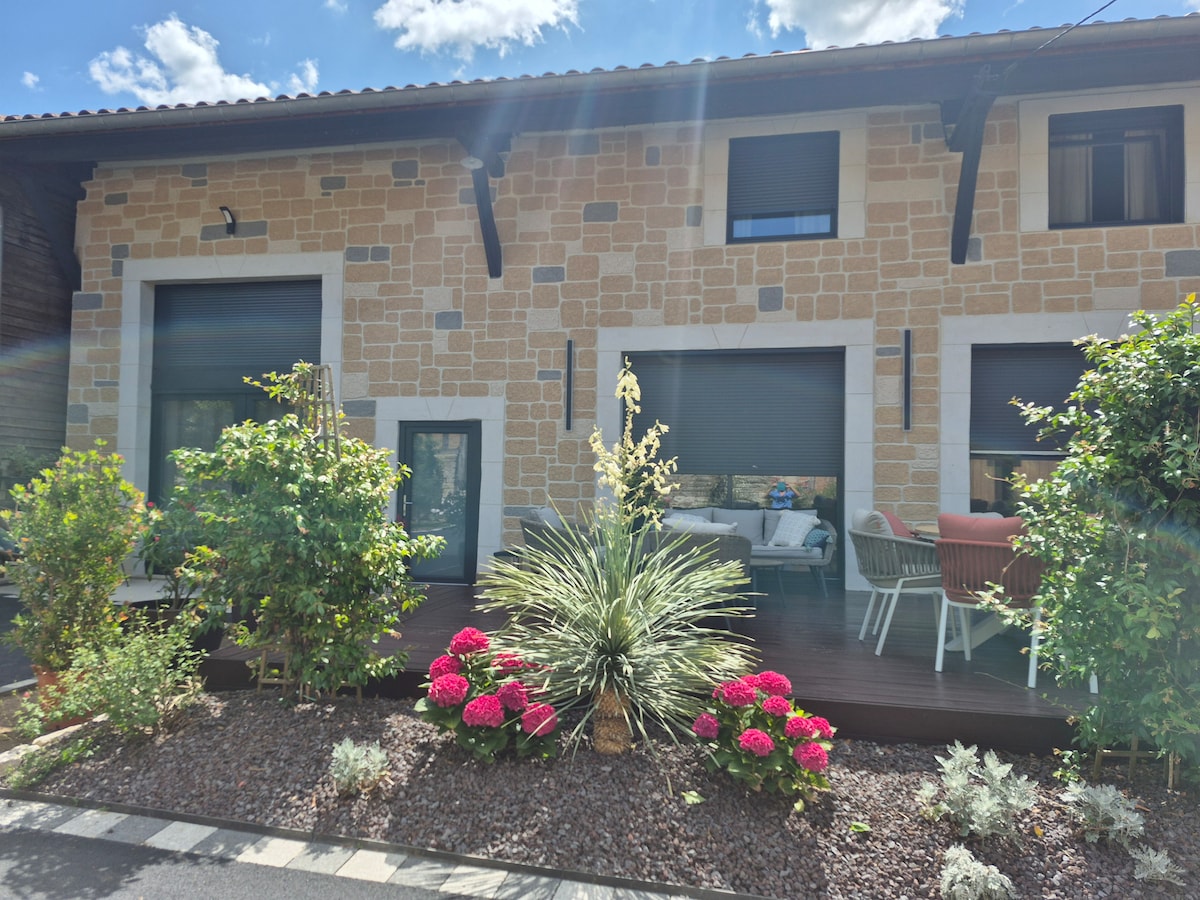 An exterior view of the property features a stone facade complemented by lush greenery and colorful flowers in the foreground. A wooden deck is visible with outdoor seating arrangements, creating a welcoming area for relaxation and socializing.