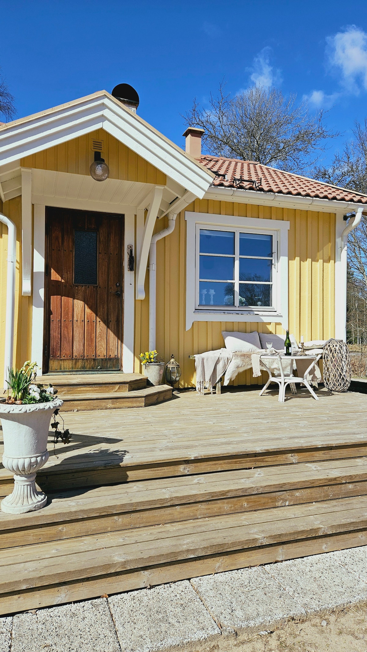 A charming yellow cottage is set against a clear blue sky, featuring a welcoming wooden door. The surrounding porch is adorned with potted plants and a small seating area, creating an inviting outdoor space for relaxation.
