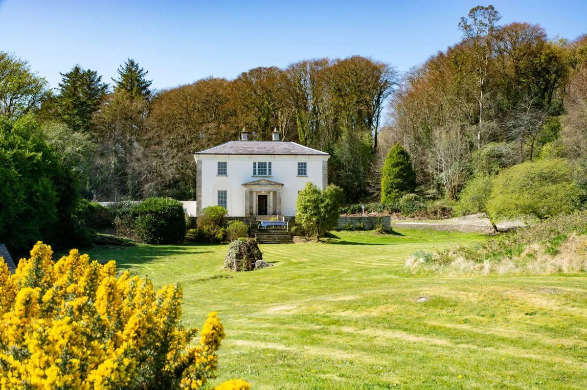 A Georgian villa is positioned amid lush greenery, with a well-maintained lawn and mature trees surrounding it. A cozy front porch is visible, complementing the home's elegant facade. Bright yellow flowers in the foreground add a vibrant touch to the serene landscape.