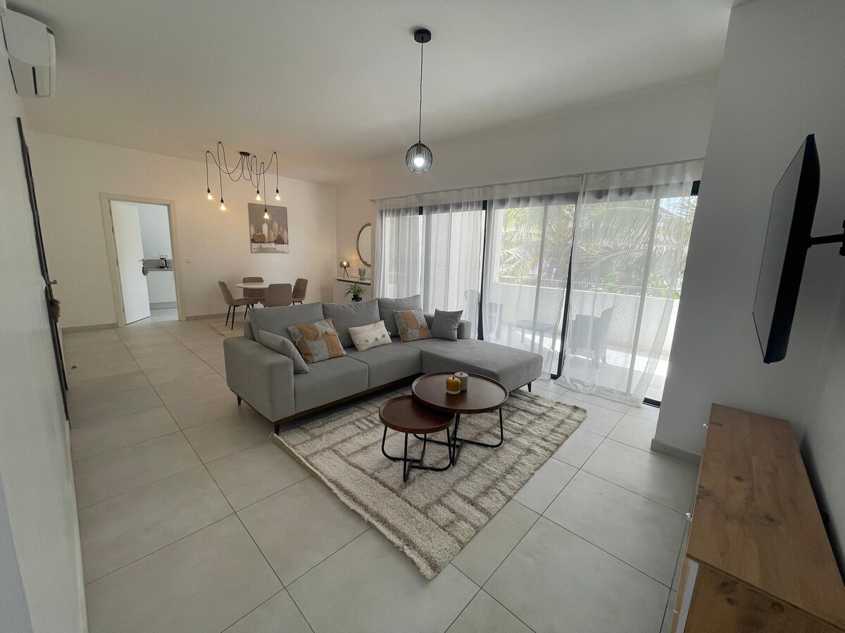 A spacious living area is portrayed, featuring a modern grey sectional sofa complemented by decorative pillows. A round coffee table sits on a textured rug, while large glass sliding doors allow natural light to fill the space, leading to a balcony adorned with plants.