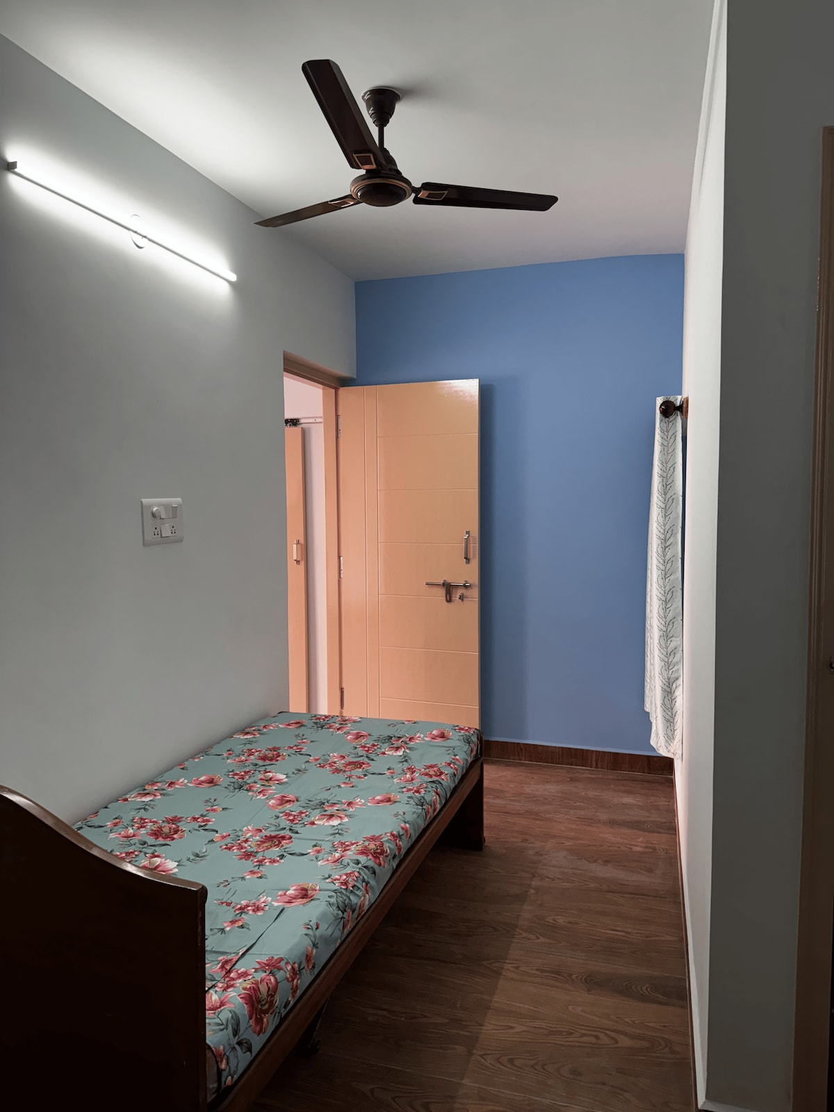 A single bed with a floral-patterned mattress is positioned against a light-colored wall. A blue accent wall adds a pop of color, while a doorway leads to a private bath. A ceiling fan is mounted above, providing airflow in the room.