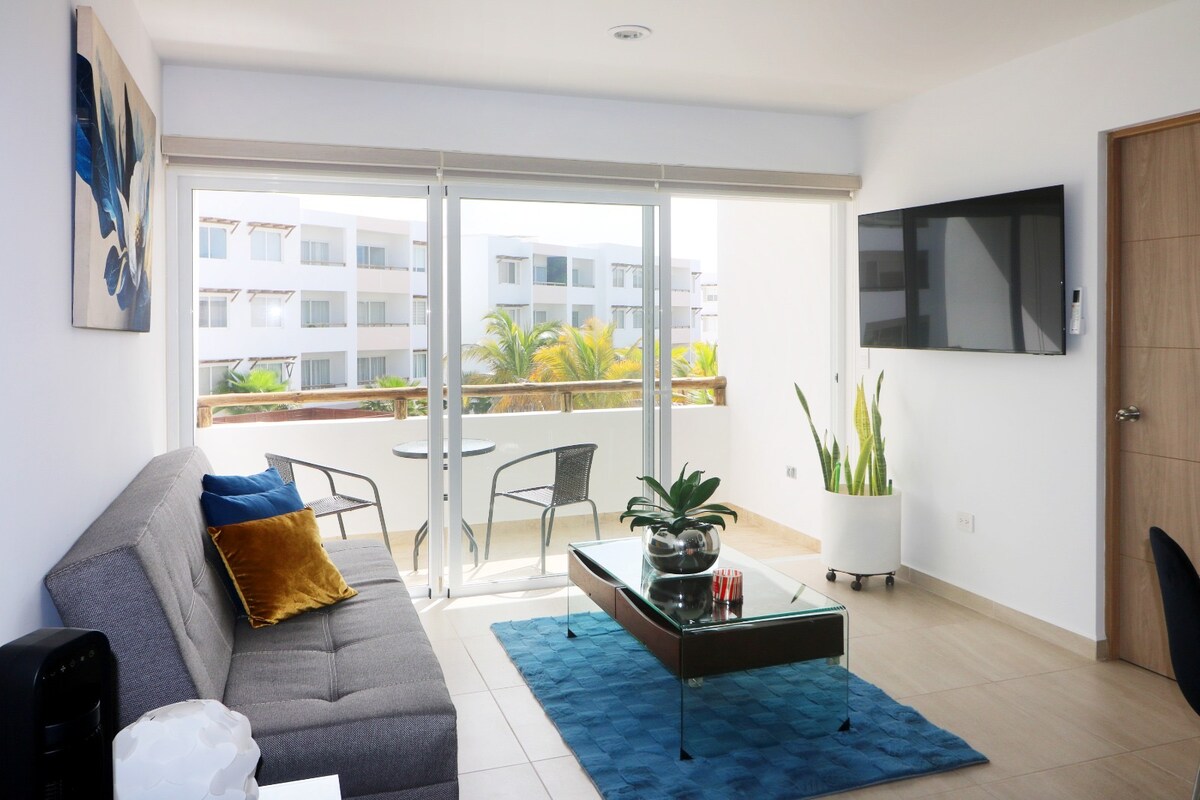 A bright living area is presented with large glass doors revealing a balcony. A gray sofa with decorative pillows is complemented by a glass coffee table and a blue area rug. Potted plants add a touch of greenery, enhancing the modern decor.