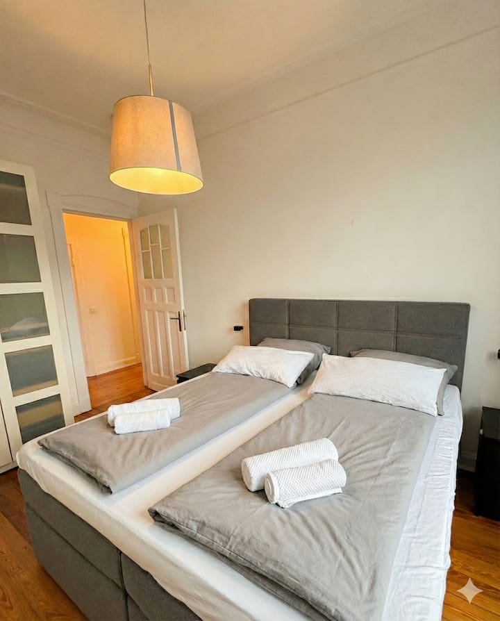 Das Zimmer bietet ein großzügiges Bett, mit frischer Bettwäsche und bereitgelegten Handtüchern für Ihren Aufenthalt. Der Raum besticht durch seinen warmen Holzfußboden, hohe Decken und ein schlichtes, ordentliches Design. Bild ist AI optimiert.