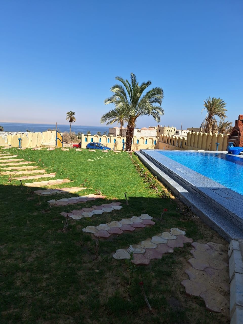 A sunlit outdoor area features a lush green lawn bordered by a pathway made of stone tiles. A swimming pool reflects the sky, with palm trees lining the space, offering a serene atmosphere near the water's edge.