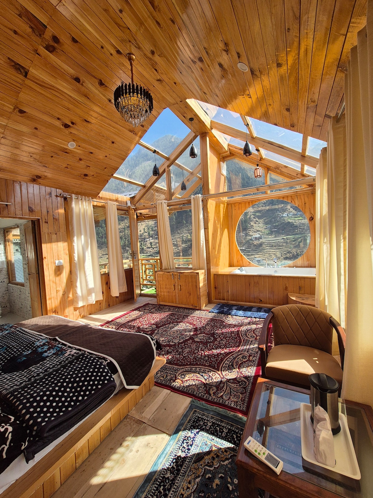 A bright and airy room features wooden walls and ceiling, accented by expansive windows allowing natural light. A spacious bed is complemented by a patterned rug. A circular window enhances the design, while a comfortable seating area includes a table and a kettle.