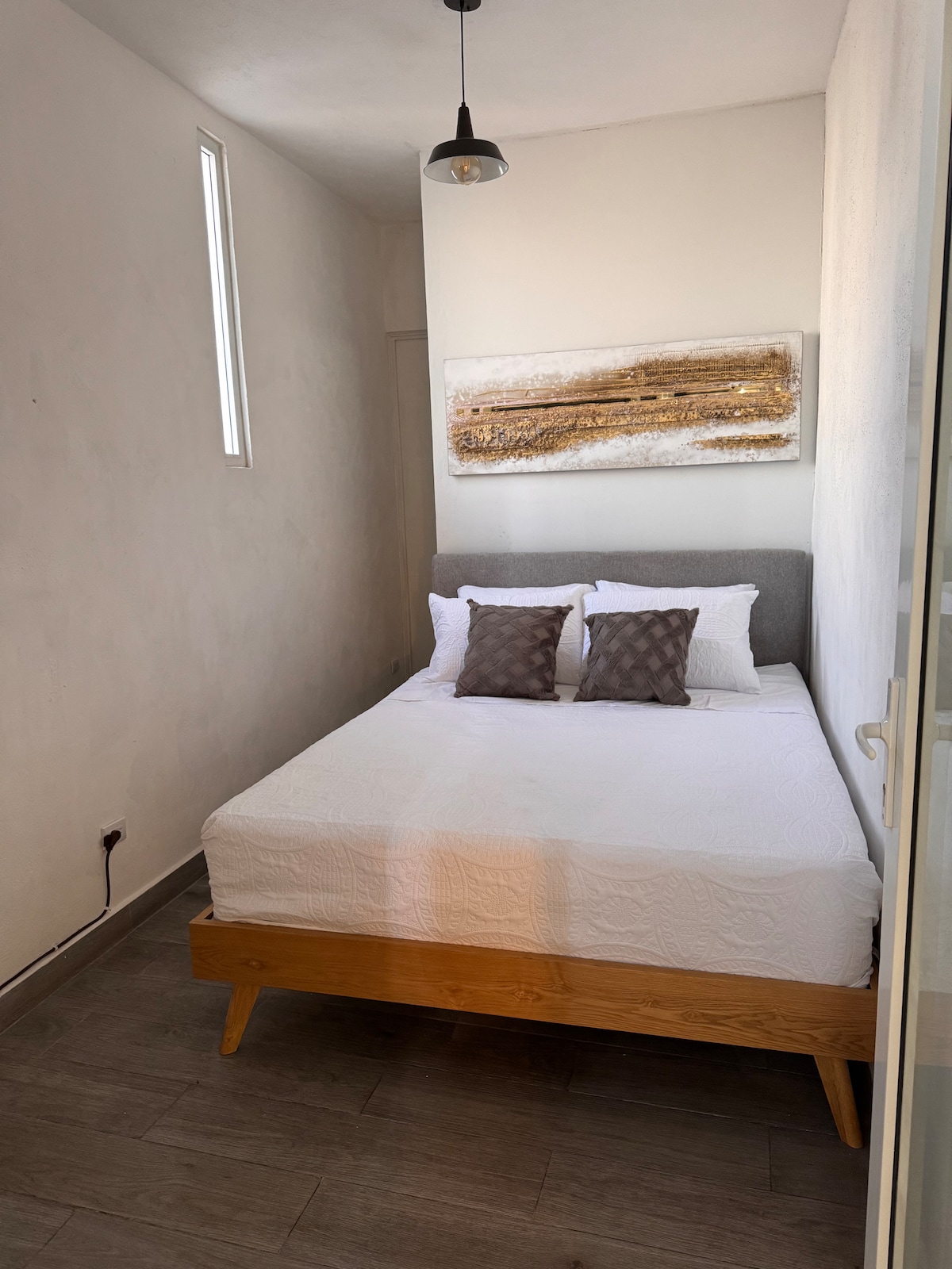 A compact bedroom features a comfortable bed with a light duvet and two decorative pillows. A piece of abstract wall art hangs above the headboard, and natural light enters through a narrow window. Simple wooden furnishings complete the functional design.