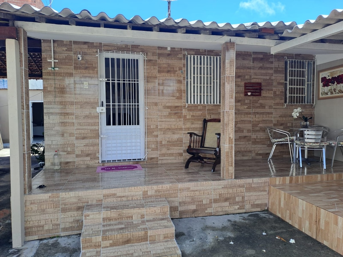 An outdoor terrace features a tiled floor and is furnished with a metal table and chairs. A wooden rocking chair is positioned nearby. The entrance is framed by stone-patterned walls and secured with a white door and window grills, providing a welcoming appearance.