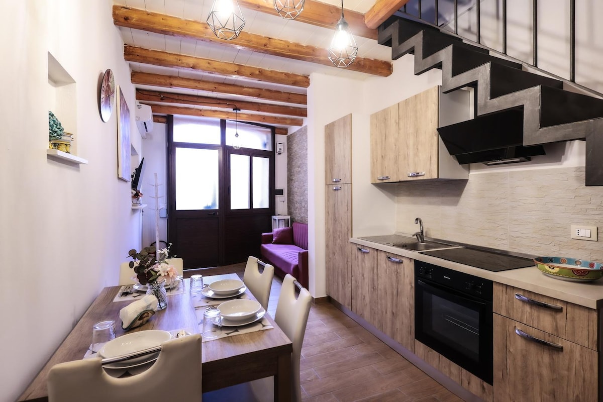 An open kitchen and dining area are visible, featuring wooden cabinetry and a modern oven. A dining table is set for four, with plates and glasses arranged neatly. Soft natural light enters through large windows, illuminating the space that connects to a cozy living area.