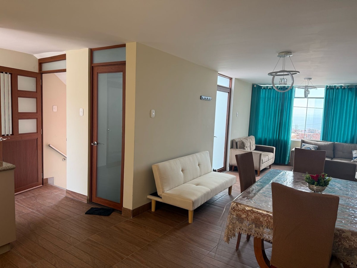 A spacious living area features elegant light-colored furnishings, including a sofa and a dining table set for six. Large windows allow natural light to fill the room, complemented by teal curtains. A staircase and a glass door are visible, leading to other areas of the apartment.