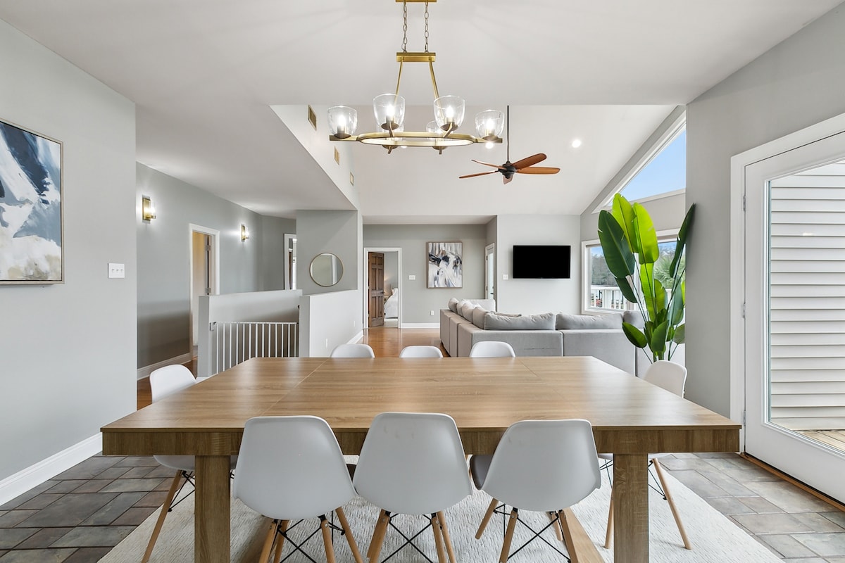 The dining area features a large wooden table surrounded by white chairs. A ceiling fan hangs above, while large windows provide natural light. The open layout connects to the living space and showcases a contemporary aesthetic with neutral tones and greenery.