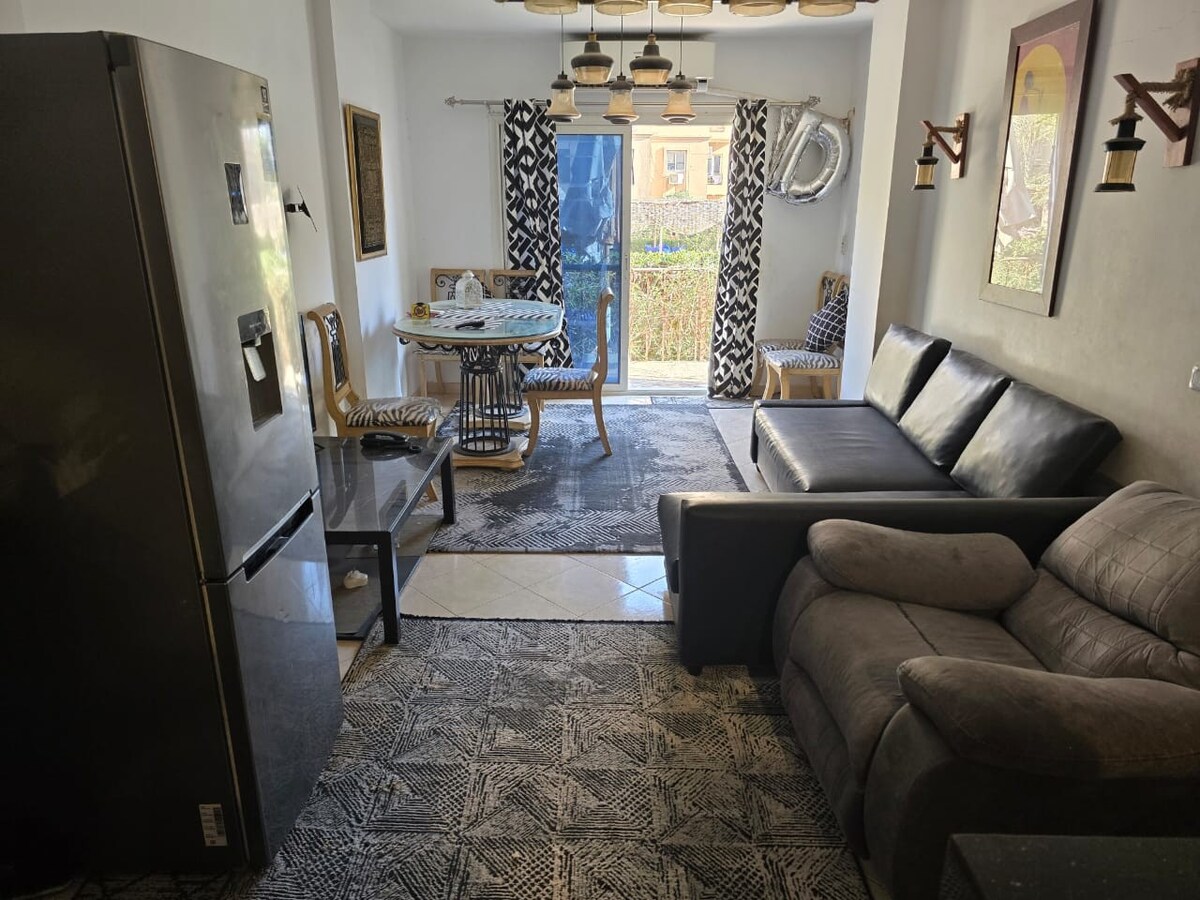 A modern living area is presented, showcasing a black leather sectional sofa and a dining table surrounded by four chairs. A large refrigerator stands in the corner. Natural light filters through glass doors, illuminating intricate floor patterns and soft textiles.