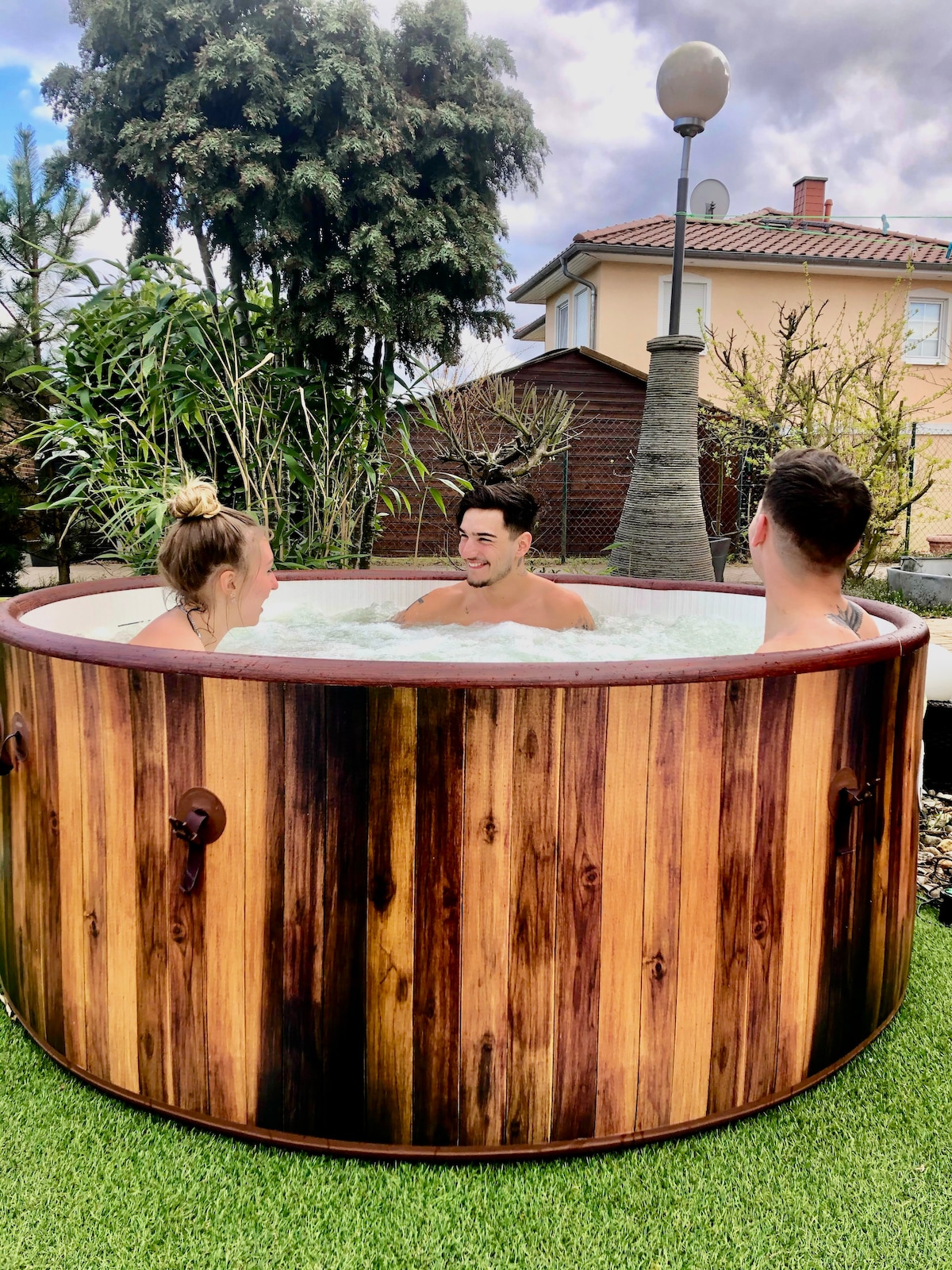 A wooden hot tub is surrounded by greenery, providing a serene outdoor setting. Three individuals are seated inside, enjoying the warm water. A soft lawn and a lamp post add to the relaxing atmosphere, enhancing the appeal for outdoor leisure.