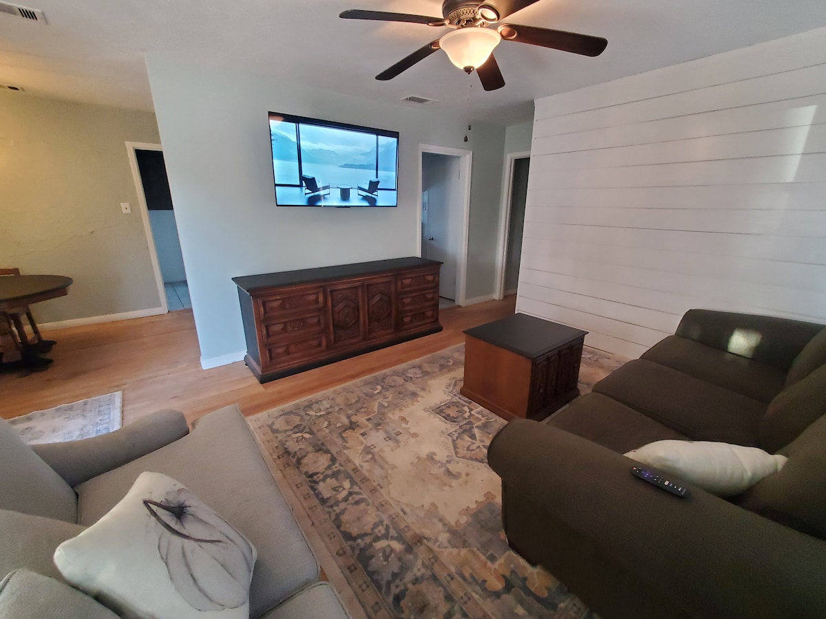 A comfortable living area features a dark wood entertainment unit with a mounted television above. A light-colored sofa alongside a darker sofa offers ample seating. A decorative area rug lies beneath a coffee table, while a ceiling fan provides airflow in the spacious room.
