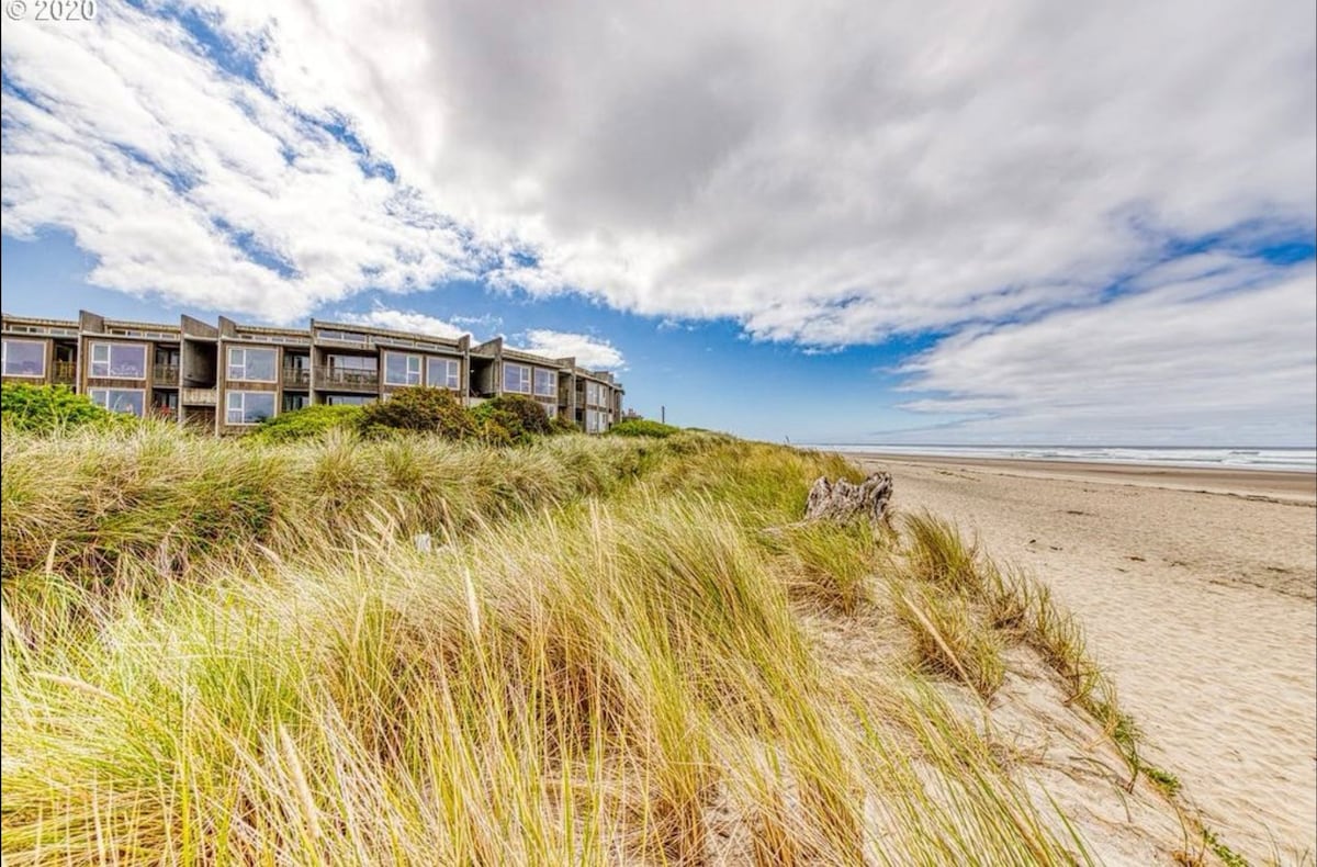A beachfront view is depicted, showcasing sandy dunes with tall grasses swaying gently in the breeze. The condo building is visible in the background against a partly cloudy sky, with hints of blue peeking through the clouds.