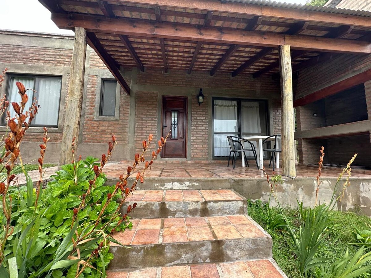 An inviting entrance features a tiled pathway leading up to a covered porch with wooden beams. A sturdy door is framed by large windows, and a small table with two chairs is positioned on the porch. Lush greenery surrounds the steps and adds a touch of nature.