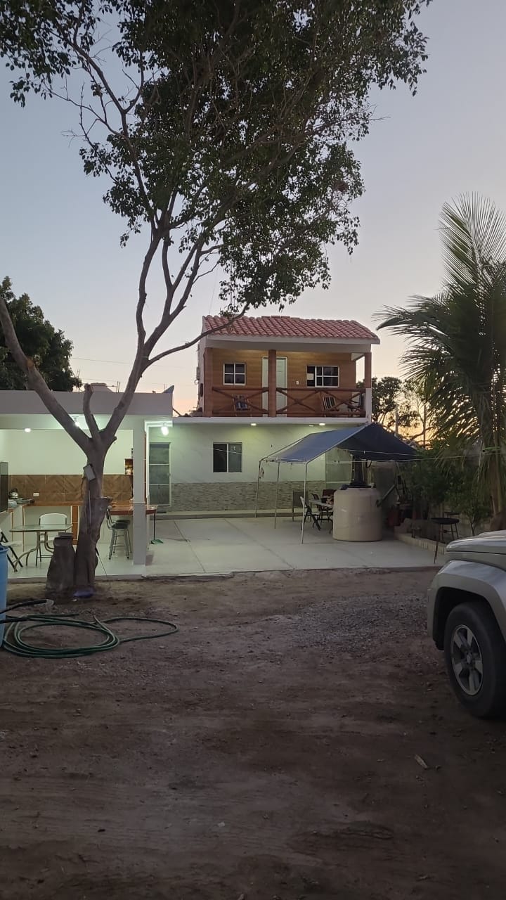 A two-story house is visible, with a wooden upper level topped by a red roof. The spacious patio area includes outdoor seating and a canopy. Lush greenery surrounds the property, while a gravel driveway leads to a parked vehicle. Soft evening light casts a warm glow.