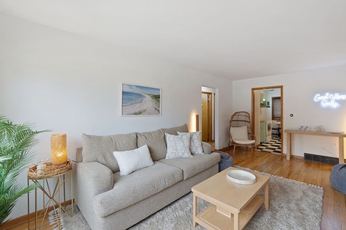 The spacious living area features a comfortable light-colored sofa adorned with decorative pillows. A cozy rug covers the wooden floor, while a small coffee table sits in front. Soft lighting is provided by a lamp, and a coastal artwork is displayed on the wall.