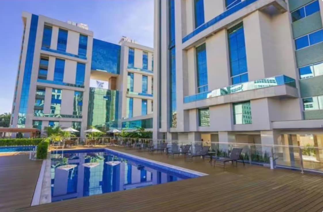 The image showcases a modern building complex reflecting blue glass facades. A serene swimming pool is framed by a wooden deck, with lounge chairs arranged nearby. Surrounding greenery and shaded areas enhance the outdoor space, creating an inviting environment.