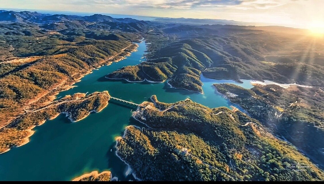 An aerial view captures a winding river surrounded by lush forests and hills. The water reflects soft colors of the sky, creating gentle contrasts. This serene landscape depicts a harmonious blend of greenery and water, emphasizing the natural beauty of the region.