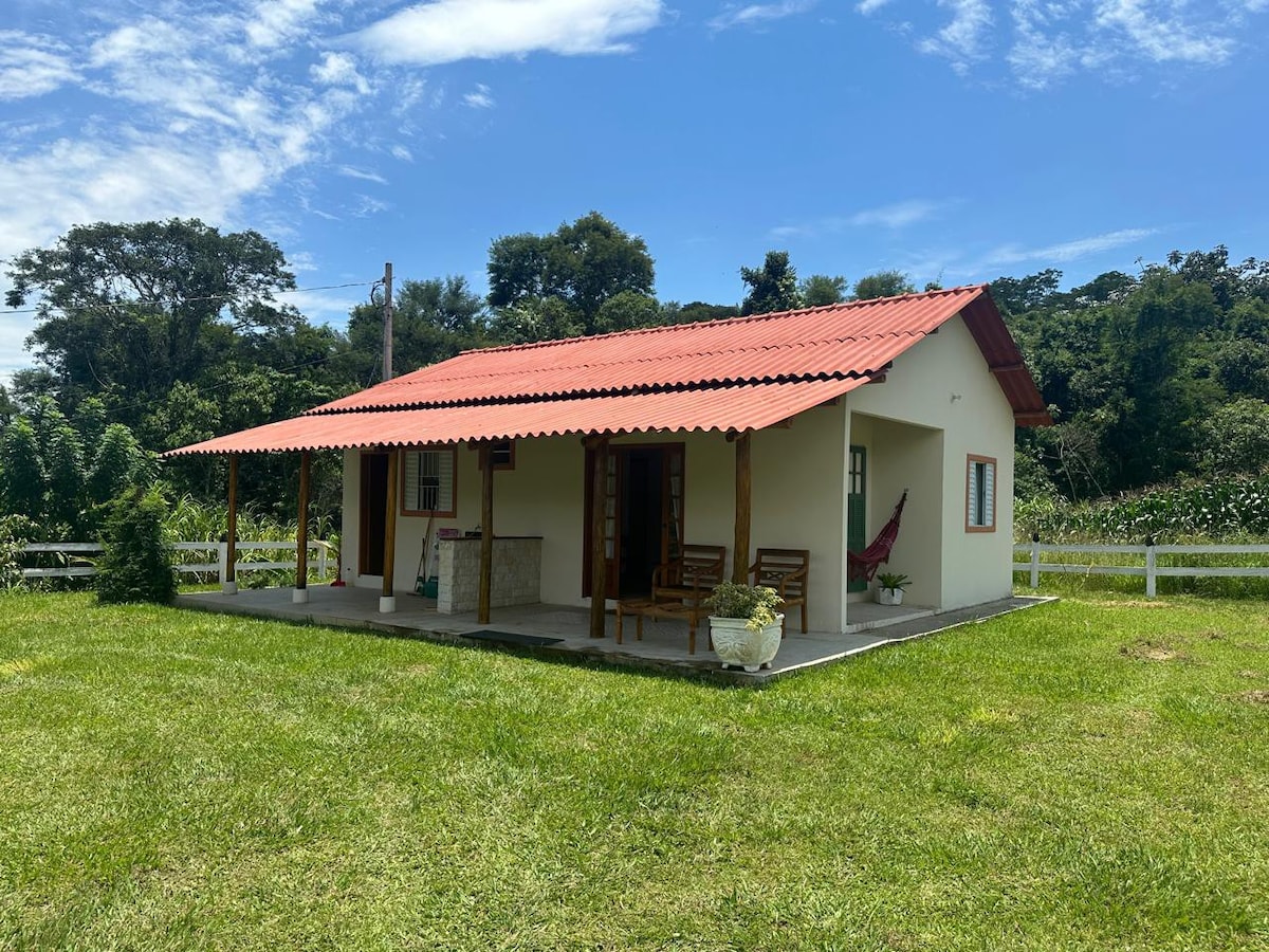 A charming rural house is set against a backdrop of lush greenery. The building features a red roof, front porch with wooden seating, and a hammock. The surrounding yard is spacious, with well-maintained grass and natural landscapes enhancing the serene environment.