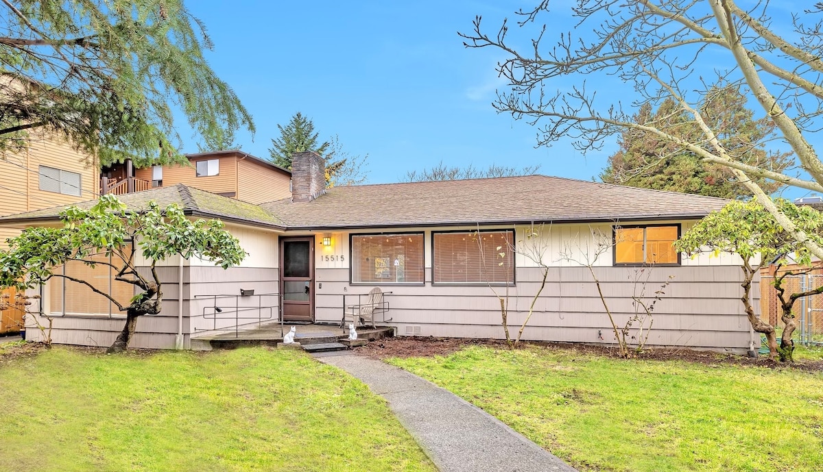 A single-story rambler is depicted, showcasing a light-colored exterior with a spacious front yard. The flat pathway leads to the entrance, framed by lush green landscaping and trees. Large windows allow natural light to fill the interior, contributing to the home's welcoming presence.