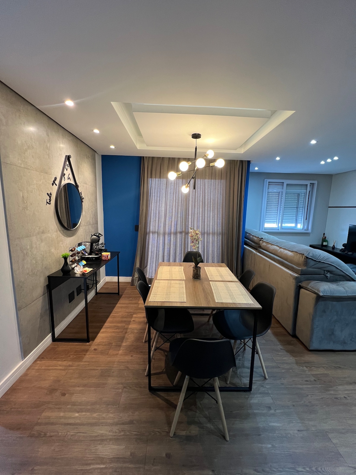 A dining area features a rectangular table surrounded by sleek black chairs. A decorative mirror is mounted on the wall, and a light fixture with multiple bulbs hangs from the ceiling. Natural light enters through curtains, illuminating the space, which connects to a cozy living area.