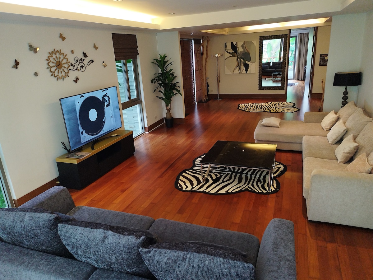 A spacious living area features wooden flooring and large windows, bringing in natural light. A plush dark grey sectional sofa faces a black television unit. A glass coffee table rests on a patterned area rug, while additional seating is arranged in the background near the sliding doors.