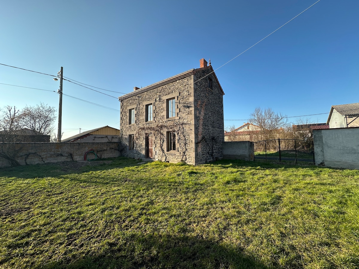 A charming stone farmhouse is depicted, showcasing its sturdy construction under clear blue skies. The grassy lawn surrounds the building, providing ample outdoor space. A fence outlines the property, and nearby structures are visible in the background, enhancing the rural atmosphere.