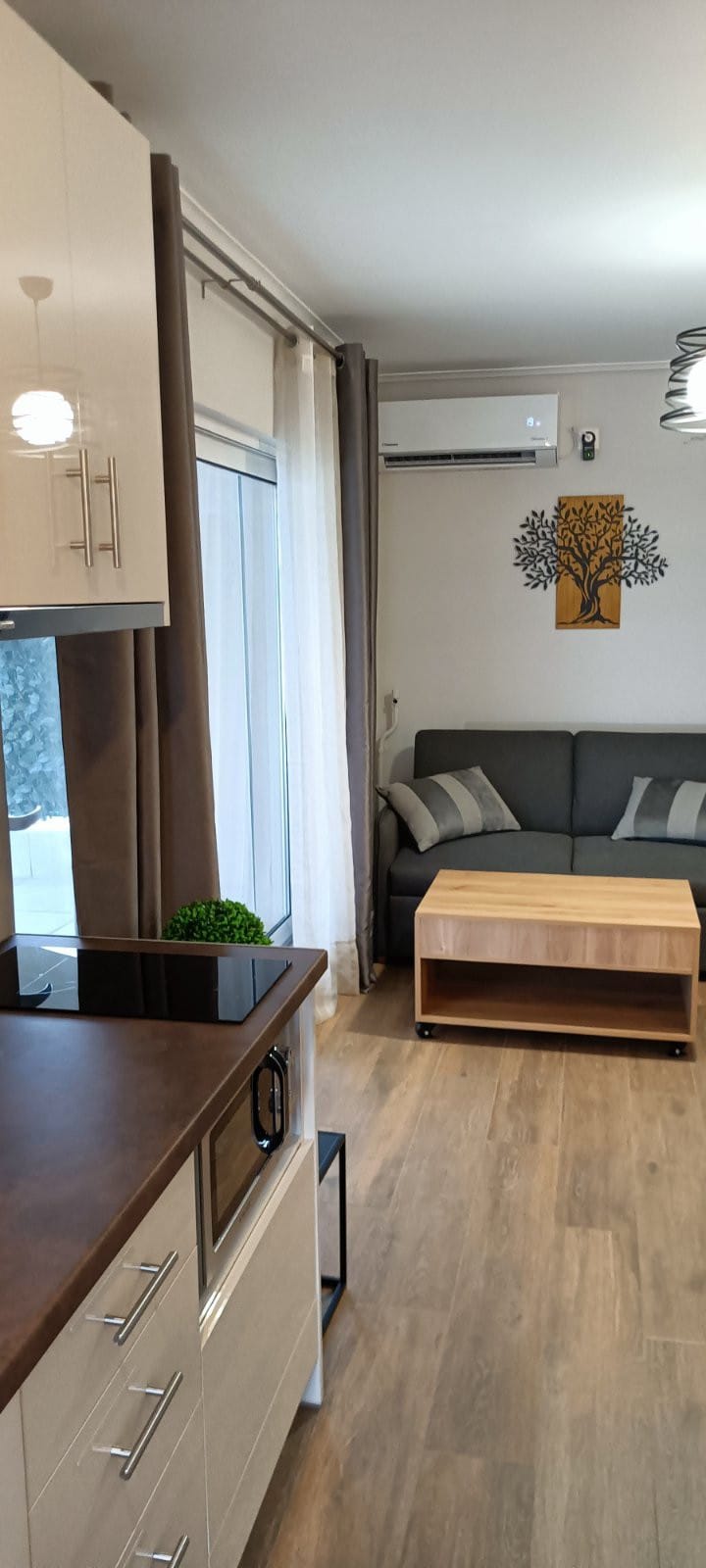 A cozy living area features a modern sofa adorned with cushions, positioned near a wooden coffee table. Natural light floods the space through large windows, dressed in sheer curtains. A decorative wall art piece of a tree complements the light-toned walls, emphasizing a calm ambiance.