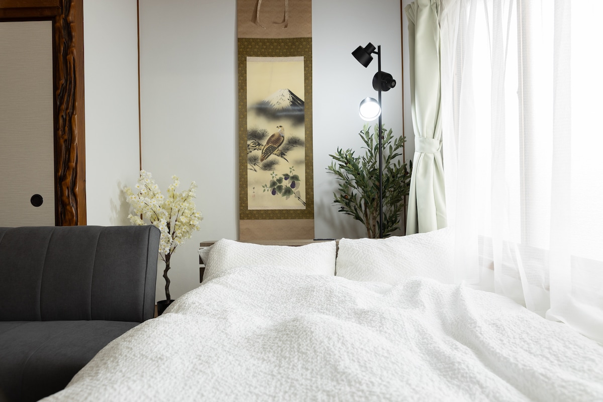 A cozy sleeping area is presented, featuring a soft white bedding set atop a spacious bed. A dark gray sofa is positioned nearby, accompanied by a decorative plant. Natural light filters through sheer curtains, illuminating a traditional wall hanging in the background.