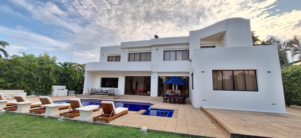 The exterior of the house is presented, showcasing a modern white facade with large windows. A pool is visible in the foreground, surrounded by lounge chairs. The patio area includes a dining table shaded by an umbrella, complemented by lush greenery in the background.