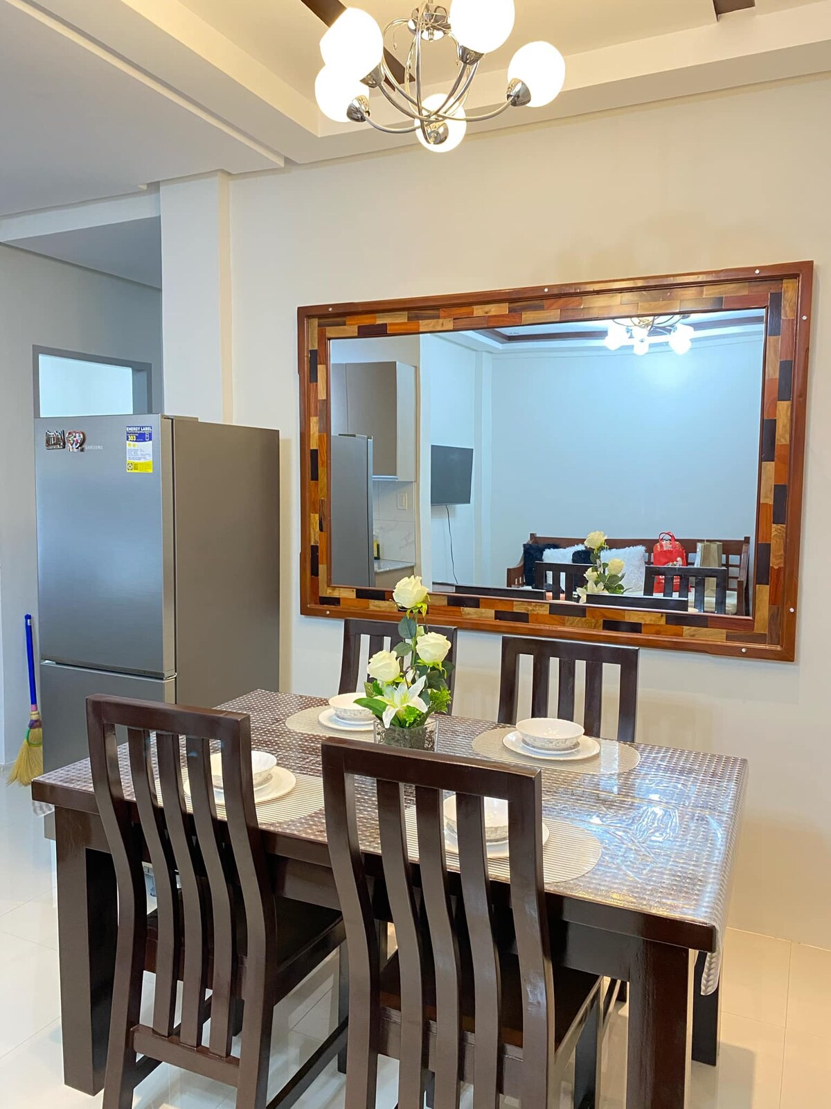 A modern dining area features a solid wood table surrounded by six matching chairs. A decorative mirror hangs above the table, reflecting the room's bright lighting and spaciousness. A stainless steel refrigerator is visible in the background, adding functionality to the inviting space.