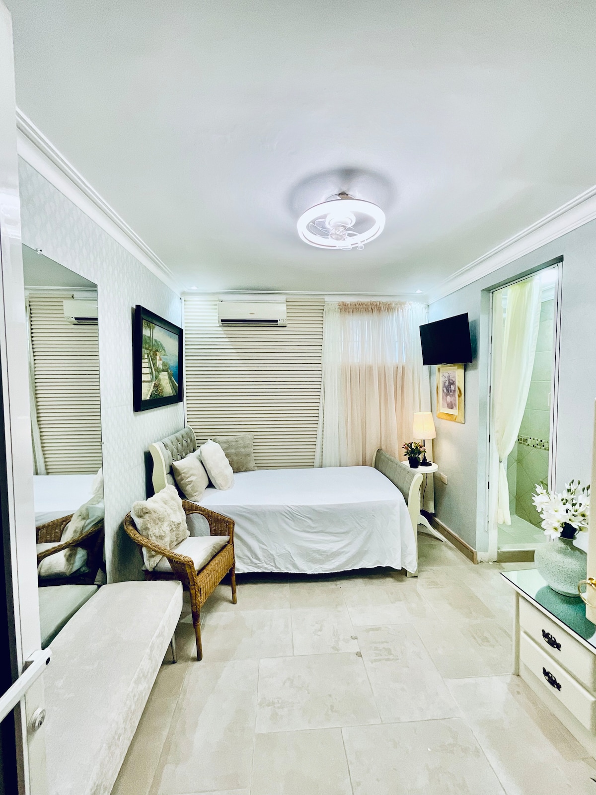 A compact bedroom is presented, featuring a spacious bed covered with a white bedspread. A wicker chair is situated beside the bed, and a small table with a lamp is positioned nearby. Light filters in through partially closed blinds, creating a soft ambiance.