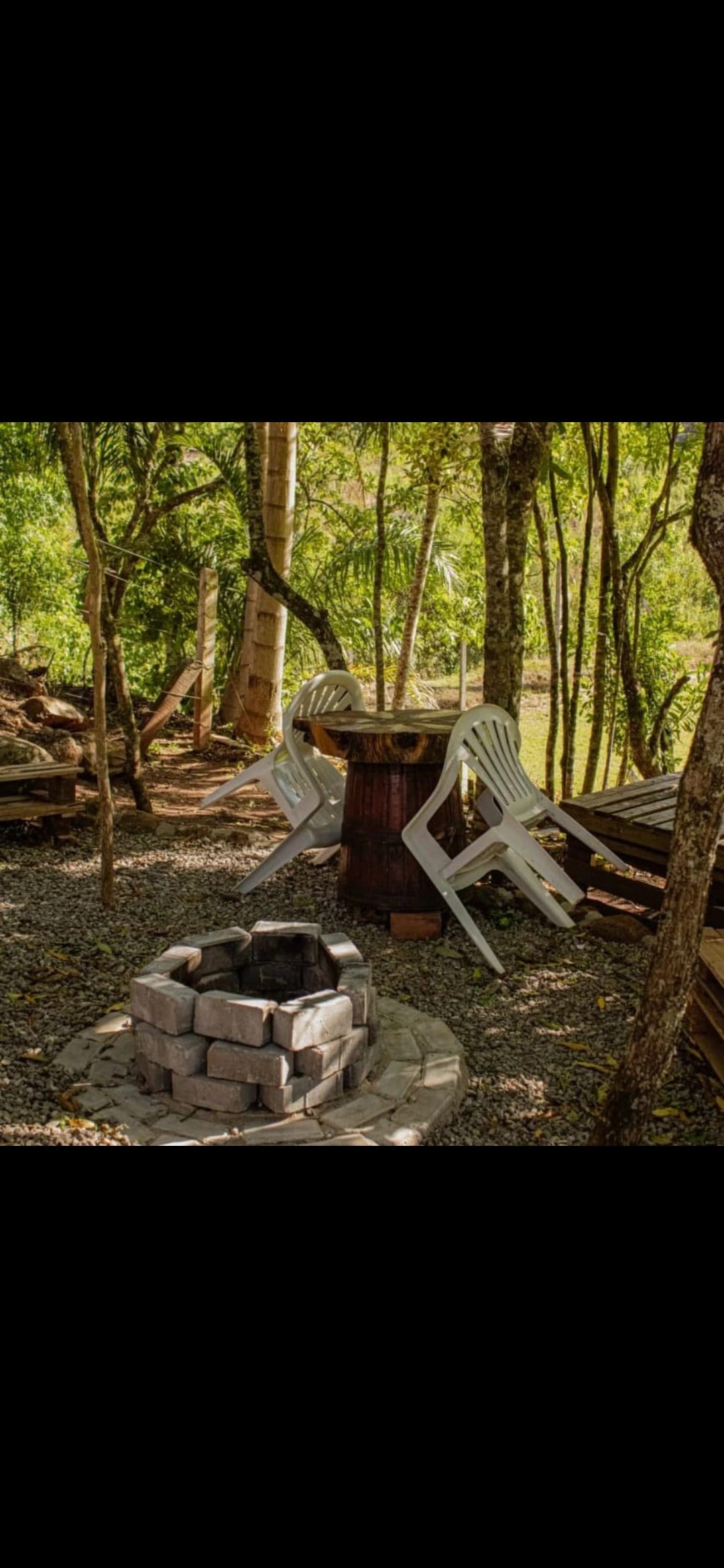 A fire pit constructed of circular gray stones is placed on a gravel surface. Two white chairs are positioned near a wooden table, surrounded by trees that provide shade. The natural setting features lush greenery in the background.